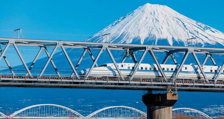 Ein Hochgeschwindigkeitszug überquert eine Brücke mit dem Berg Fuji im Hintergrund.