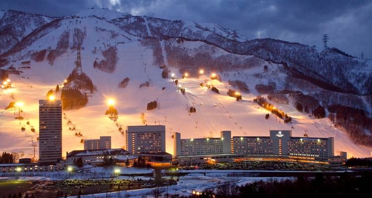 Abendlicher Blick auf ein Skigebiet mit beleuchteten Gebaeuden und Sesselbahnlinien an einem verschneiten Berghang unter einem daemmernden Himmel.