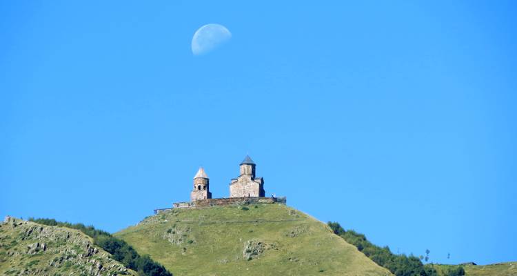 Église sur une colline avec une lune visible dans le ciel clair.