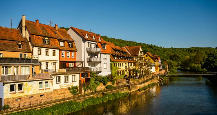 Fila de casas históricas coloridas que bordean un río tranquilo con colinas boscosas al fondo en un día soleado y brillante.
