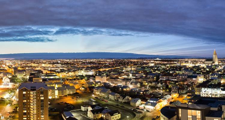 Panoramisch avondaussicht over Reykjavik met glinsterende stadslichten en de opvallende Hallgrimskirkja kerk onder een dramatische schemering.