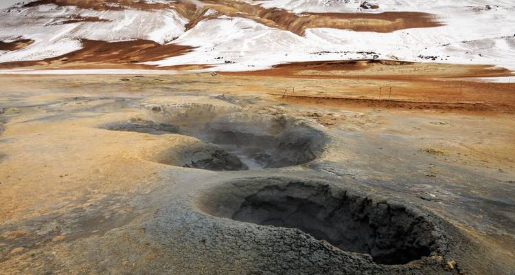 Stoomende geothermische modderpotten tegen een kaal, sneeuwvlekkerig IJslands bergachtig landschap.