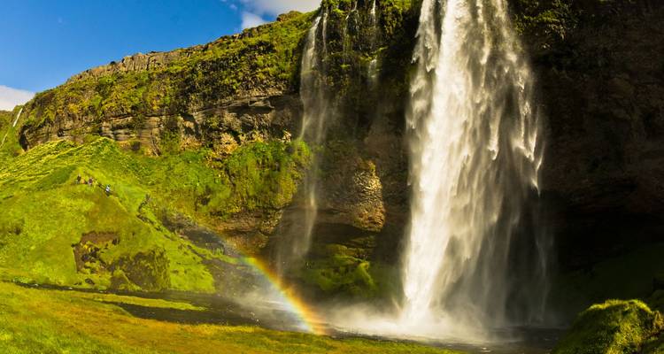 Een indrukwekkende waterval die over een bemoste klif naar beneden stort met een levendige regenboog in de mist en kleine wandelaars in de buurt.
