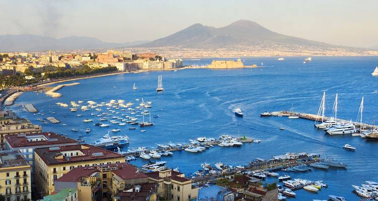 Uitgestrekt uitzicht over de waterkant van Napels vol met bootjes, met de Vesuvius die uit de baai oprijst.