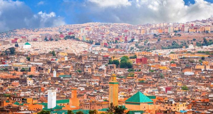 Expansive view over Fes medina with colorful rooftops and distant hills under partly cloudy skies.