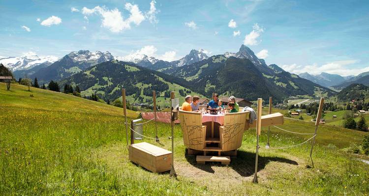 Eine Familie genießt ein Picknick im Freien an einem Holztisch in der Form einer Milchkanne vor einer panoramischen Alpenwiese.