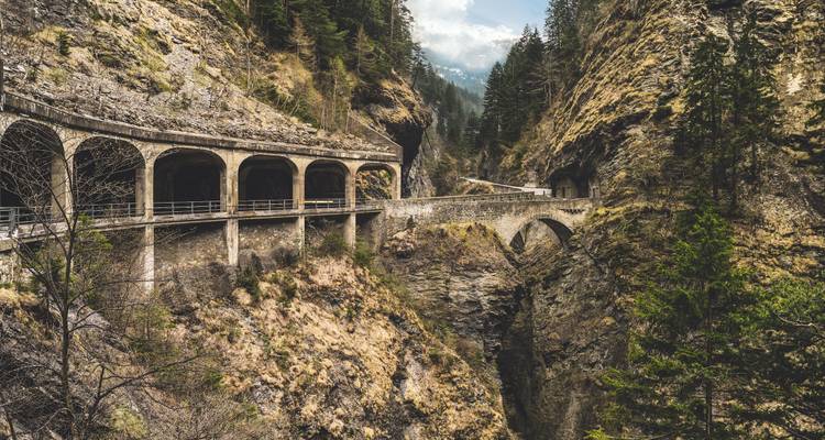 Historische Bogenbauwerke und Brücke klammern sich an steilen Felswänden über einer beeindruckenden Bergschlucht.