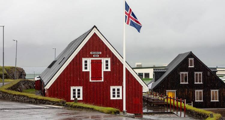 Traditioneel rood houten IJslands huis met nationale vlag in licht regen tegen een grijze kustachtergrond.