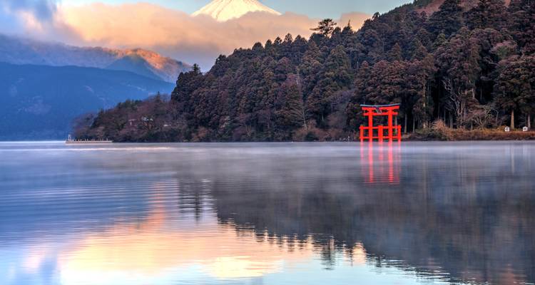 Vista neblinosa del amanecer sobre el Lago Ashi con el Monte Fuji distante, agua espejada y puerta torii roja reflejándose.