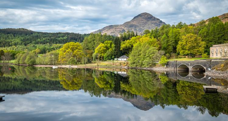 Glass-still Loch Lomond mirrors wooded slopes and arched bridge beneath rolling hill