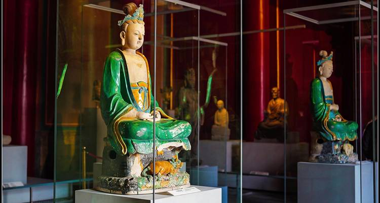 Oude keramische Boeddha-beelden tentoongesteld in glazen vitrines in een halfdonker museum.