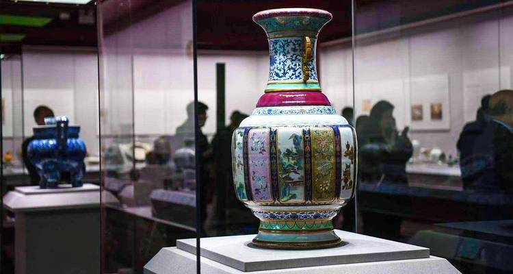 Large elaborately decorated porcelain vase displayed in a well–lit glass case inside a museum gallery, with silhouettes of visitors observing exhibits in the background.