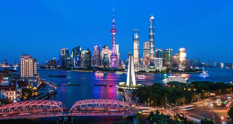 Illuminated Shanghai skyline at dusk featuring the Oriental Pearl Tower, Shanghai Tower and colourful reflections in the Huangpu River with boats and bridges in view.