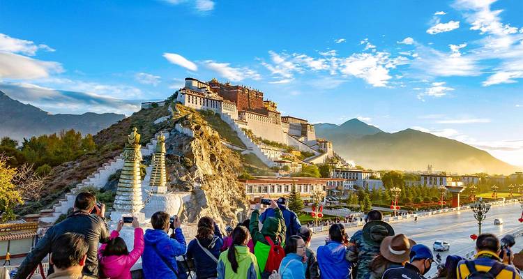 Crowd of visitors photographing the iconic Potala Palace perched on a rocky hill under bright blue skies with mountains in the distance.