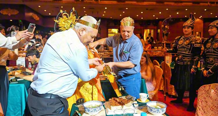 Lively indoor banquet with guests wearing festive crowns carving roast meat.