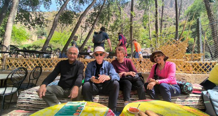 Quatre voyageurs souriants sur des coussins à motifs dans un café extérieur ombragé avec de grands arbres au-dessus.