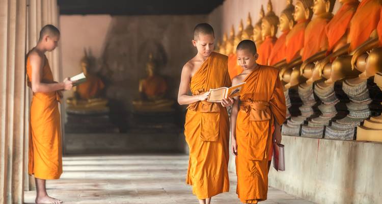 Mönche in orangefarbenen Gewändern, die in einem Tempel mit Buddha-Statuen stehen und lesen.