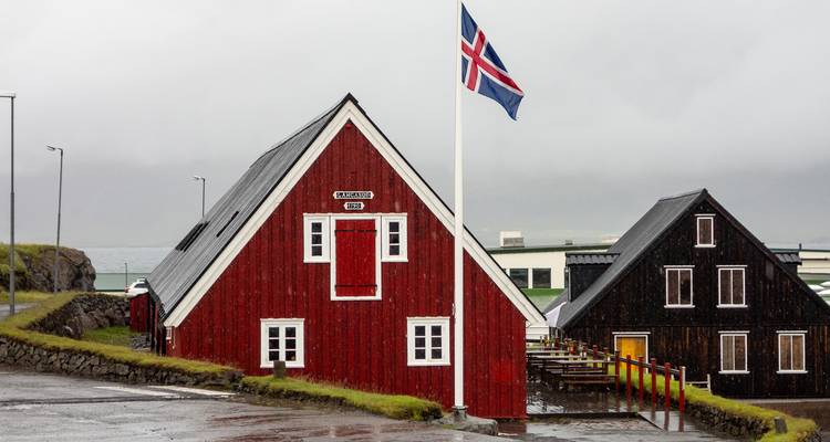 Maison en bois rouge avec drapeau islandais qui flotte sous la pluie côtière brumeuse à côté d'un bâtiment vêtu de noir.