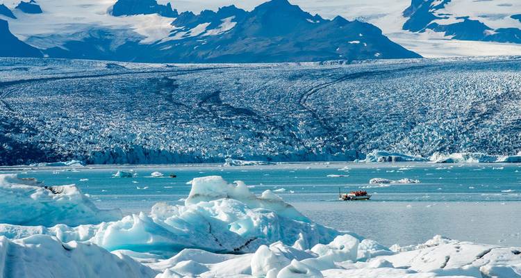 Lagon glaciaire parsemé d'icebergs bleus et d'un petit bateau de visite naviguant sous des montagnes glaciaires abruptes.