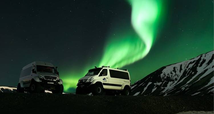 Deux super-jeeps robustes garés sous les aurores boréales d'un vert éclatant au-dessus d'une crête islandaise enneigée la nuit.