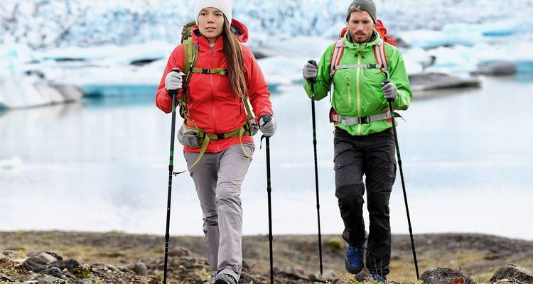 Zwei Wanderer in leuchtenden Jacken nutzen Stöcke beim Wandern neben einer vereisten Lagune und einem Gletscher in Island.