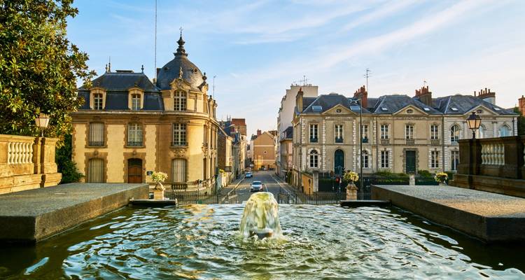 Historic city street flanked by ornate buildings and a water fountain.