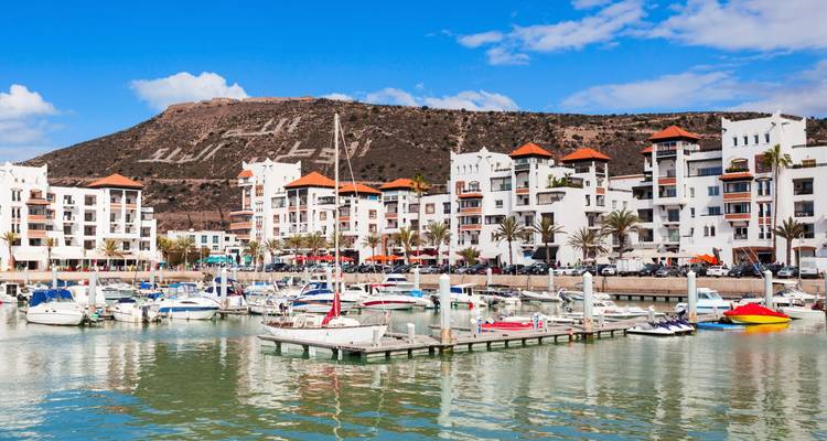 Küstenstädtchen mit weißen Gebäuden, Booten im Wasser und Hügeln im Hintergrund.