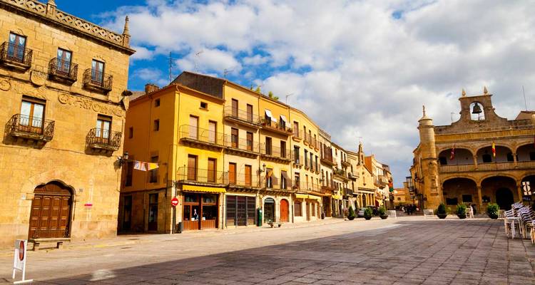 Spanish town plaza with colorful buildings under a cloudy sky.