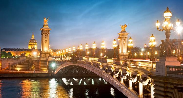 Beleuchtete Brücke über dem Fluss mit historischen Gebäuden im Hintergrund bei Nacht.