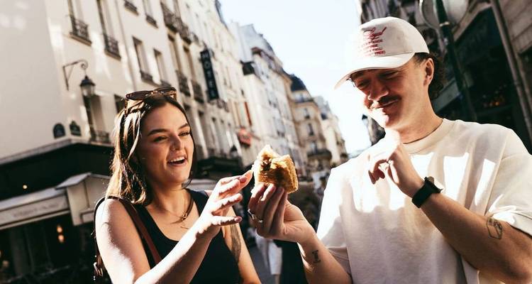 Two friends share a pastry on a lively European street lined with cafés and historic buildings.
