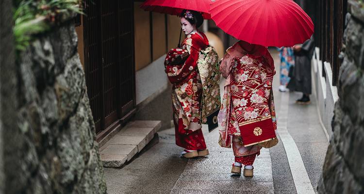 Deux personnes en kimonos traditionnels sous des parapluies rouges dans une rue étroite.