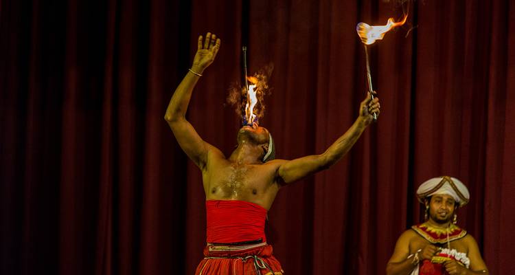Ontblote performer ademt vuur op het podium, vlammen springen op tegen dieprode gordijnen terwijl drummer toekijkt.