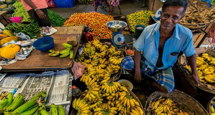 Marktverkoper omringd door stapels heldergele bananen en kleurrijke producten in een drukke Sri Lankaanse kraam.