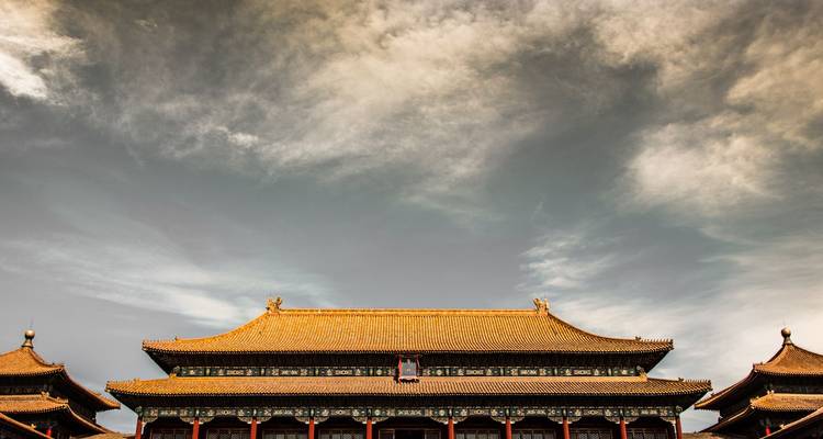 Golden-roofed hall of the Forbidden City beneath a dramatic cloudy sky, captured symmetrically.