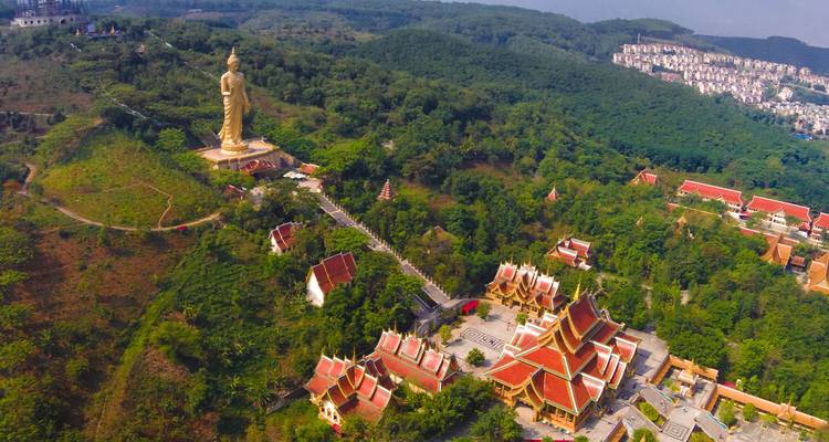 Vue aérienne d'une statue de Bouddha doré au sommet d'une colline dominant un complexe de temples aux toits rouges entouré de végétation luxuriante.