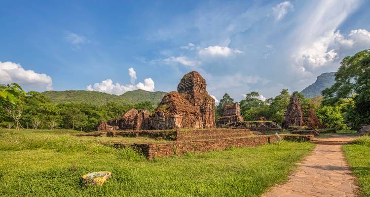 Red-brick temple ruins of Mỹ Sơn Sanctuary sit amid lush greenery and distant mountains under blue skies.