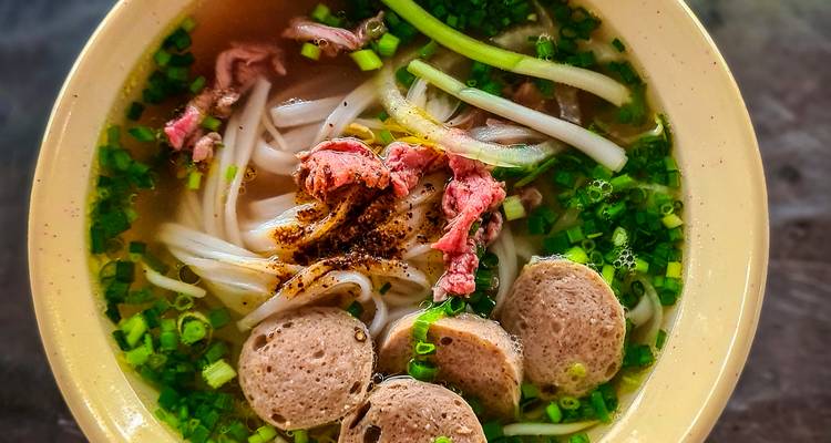 Top-down view of a steaming bowl of Vietnamese pho with beef slices, meatballs and fresh herbs.