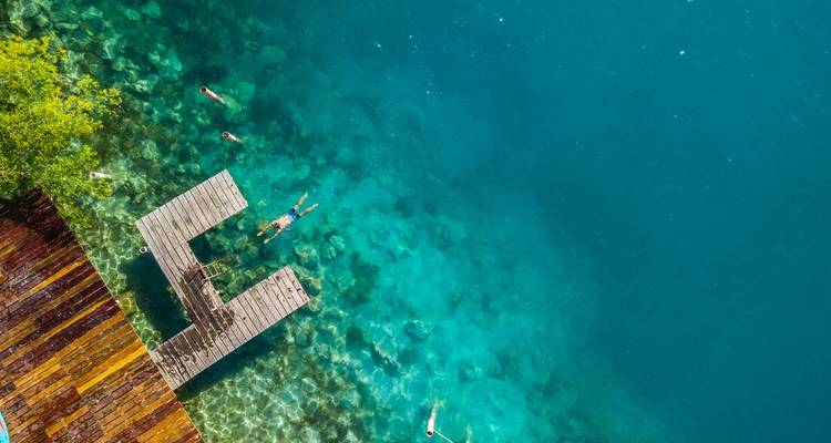 Vue aérienne de personnes nageant près d'un quai en bois au-dessus d'une eau bleue claire.