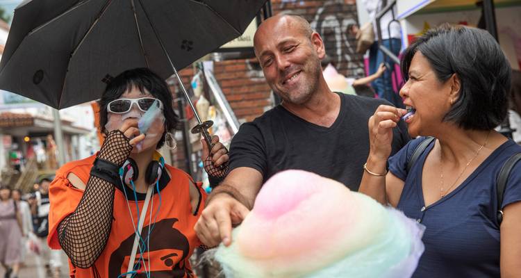 Unos amigos comparten algodón de azúcar de colores pastel mientras se resguardan bajo un paraguas en una calle muy concurrida.