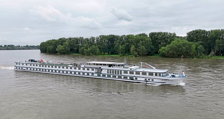 Large river cruise ship sailing through a river with trees.