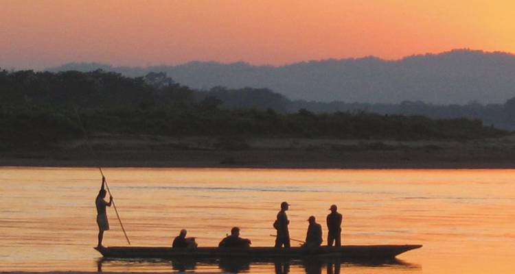 Silhouettes of several people pole a wooden boat across a calm river at dusk.
