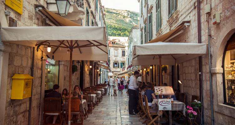 Callejón estrecho de piedra con cafés al aire libre donde los comensales están sentados bajo sombrillas y las montañas se elevan al fondo.