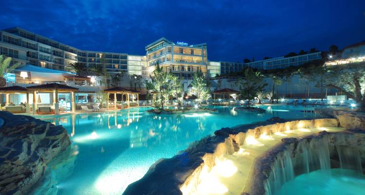 Hotel de lujo junto al mar con amplias piscinas estilo laguna iluminadas en turquesa bajo un profundo cielo nocturno azul.
