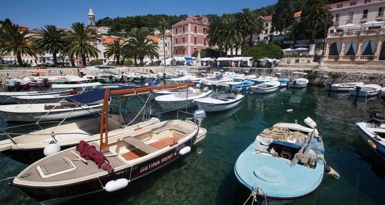 Pequeñas barcas se mecen en un puerto de agua turquesa cristalina bordeado de palmeras y edificios mediterráneos en tonos pastel.