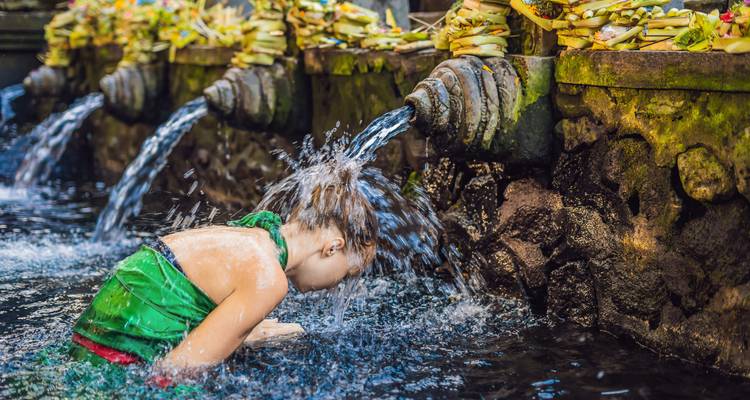 Junge Frau in grünem Sarong, die sich unter einem steinernen Ausguss verbeugt, während heiliges Quellwasser über ihren Kopf fließt in einem balinesischen Tempelbecken, das mit moosbedeckten Schnitzereien gesäumt ist.