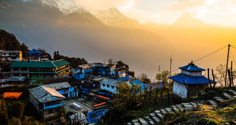 Un village de montagne brille dans la douce lumière du lever de soleil avec les silhouettes brumeuses de l'Himalaya au loin.