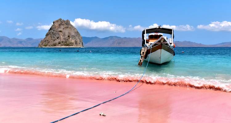 Traditionelles Holzboot vor Anker vor einem beeindruckenden rosafarbenen Sandstrand mit türkisfarbenem Wasser und fernen zerklüfteten Inseln.