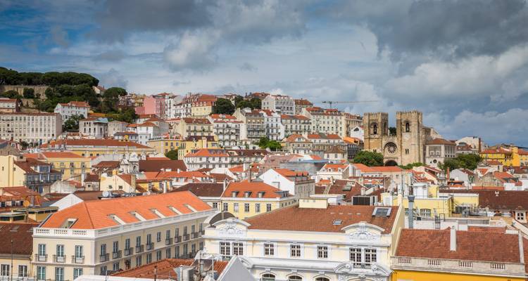 Panoramic view over the red-tiled rooftops of Lisbon with the twin towers of the Sé Cathedral under a partly cloudy blue sky.
