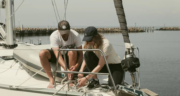 Dos viajeros trabajan juntos amarrando cuerdas en la proa de un velero en un puerto tranquilo.