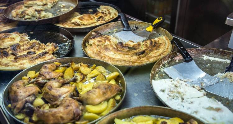 Sélection de plats balkaniques tels que des pâtisseries cuites au four, du poulet rôti et des pommes de terre présentés dans des récipients métalliques derrière un comptoir vitré.
